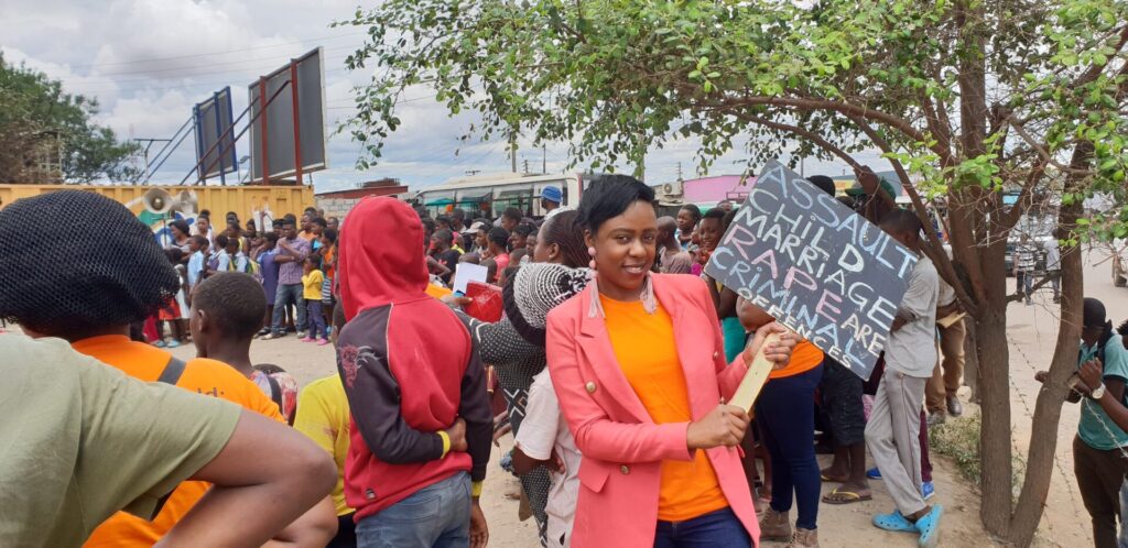 An African woman with short black hair is holding a sign saying 'assault, child marriage, rape are criminal offences'. She is part of larger crowd. 