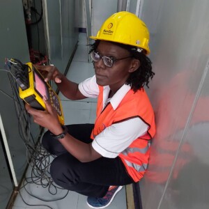 a black woman in a yello helmet and orange vest is crouched looking at a measuring device
