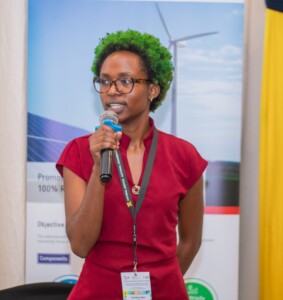 An african woman with green hair and glasses is wearing a red dress and speaking into a microphone