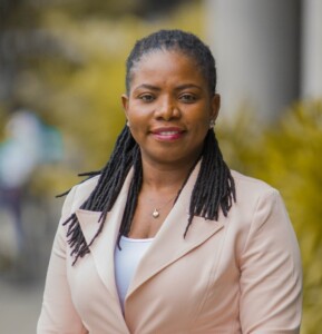 A black woman with braids, wearing a beige jacket.
