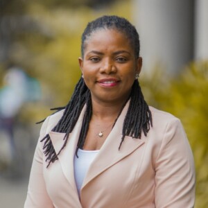 A black woman with braids, wearing a beige jacket.