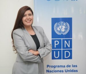 A smiling woman with long brown hair, wearing light grey suit stands next to a blue sign which says PNUD.